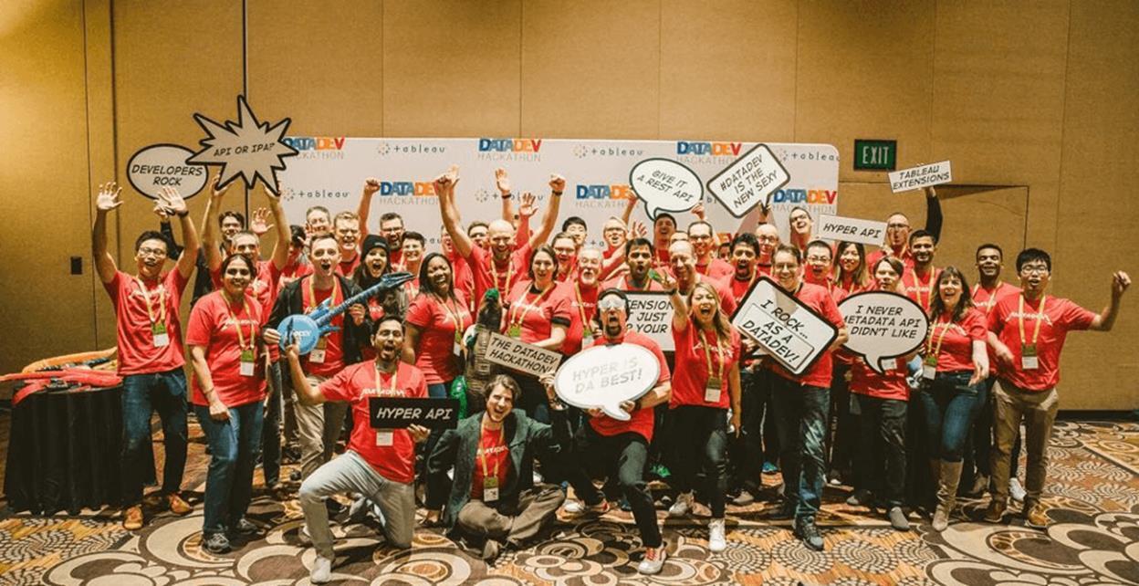 Large group cheering and smiling, all wearing red TC t-shirts and holding fun data and dev-themed signs, posing in a conference room