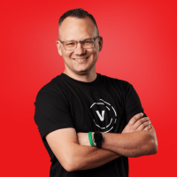 Sean is looking towards the camera smiling with his arms crossed, wearing a black Visionary t-shirt, posed in front of a bright red backround